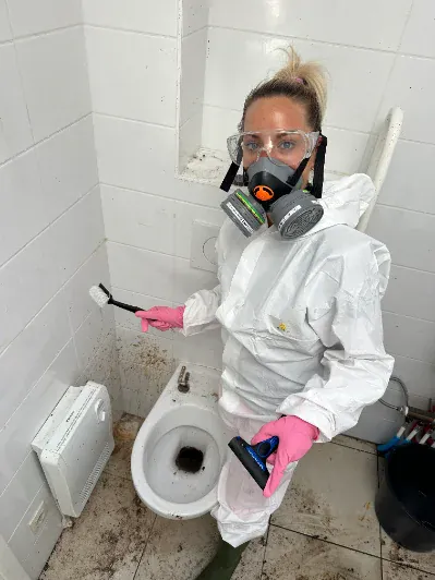 Une intervenante MelaClean en combinaison et masque, brosse à la main, dans une salle de bain en cours de nettoyage